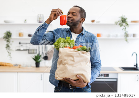 An African American man holding a bag of groceries and smelling a red bell pepper in a bright kitchen setting. An African American man holding a bag of groceries and smelling a red bell pepper in a bright kitchen setting. 133499017