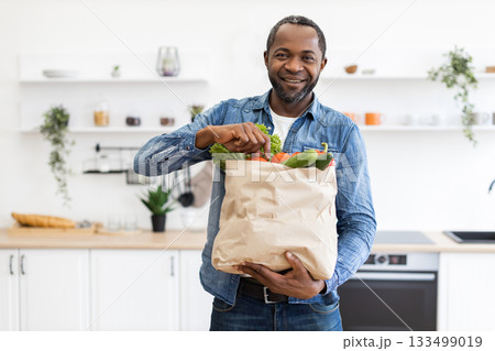 A smiling Black man holds a paper bag filled with fresh vegetables in a bright, modern kitchen. A smiling Black man holds a paper bag filled with fresh vegetables in a bright, modern kitchen. 133499019