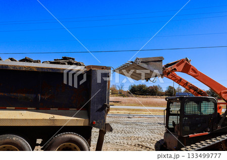 Skid steer loader unloads soil from pile into dump truck at construction site. 133499777