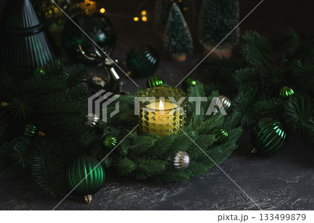 Lit Candle in Green Glass Holder with Pine Wreath and Christmas Ornaments 133499879