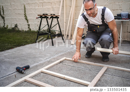 Mature male carpenter using electric screwdriver on a wooden plank outdoors 133501435