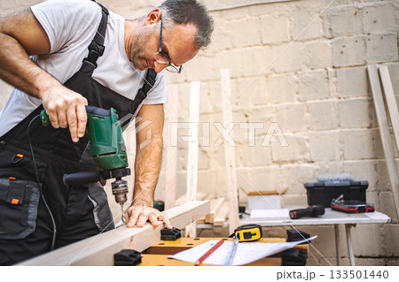 Mature male carpenter using electric screwdriver on a wooden plank outdoors 133501440