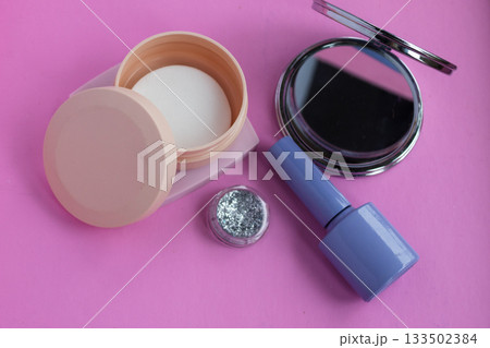 Cosmetics arrangement on a vibrant pink background featuring a compact mirror, loose glitter, a container of nail polish remover wipes, and nail polish, symbolizing beauty and self care 133502384