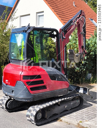 A compact mini excavator on a crawler track against the backdrop of a private residential building A compact mini excavator on a crawler track against the backdrop of a private residential building 133503465