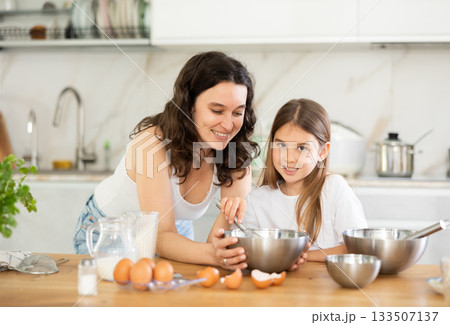 Mom and girl are talking in kitchen and mixing ingredients for pastry in bowl with whisk 133507137