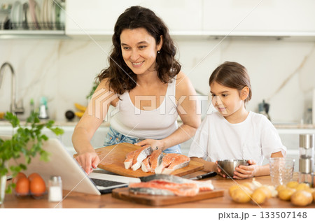 While cooking salmon fillet fish dish, woman and little daughter look at laptop 133507148