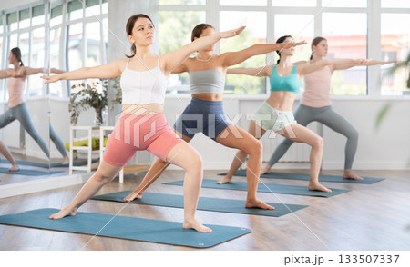 Young girl teaches yoga lesson, shows students technique of performing warrior pose asana Young girl teaches yoga lesson, shows students technique of performing warrior pose asana 133507337