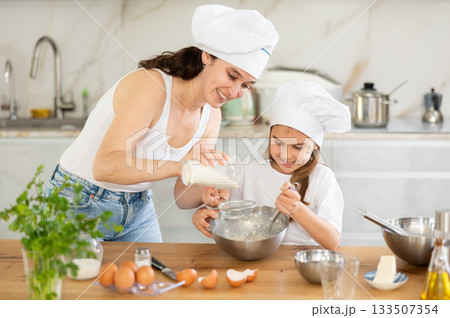 Mom sifts and adds flour to bowl, girl mixes ingredients with whisk. Mom sifts and adds flour to bowl, girl mixes ingredients with whisk. 133507354