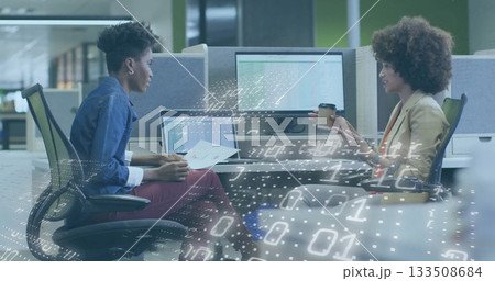 Two colleagues reviewing documents on silver laptop and holding coffee cup in office, with monitors 133508684