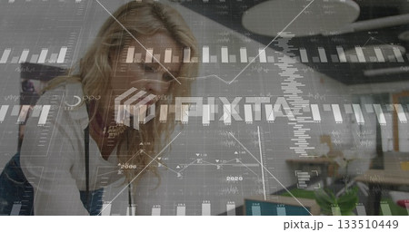 Leaning data analyst reviewing charts on laptop at open plan office desk, with notebook, plant 133510449