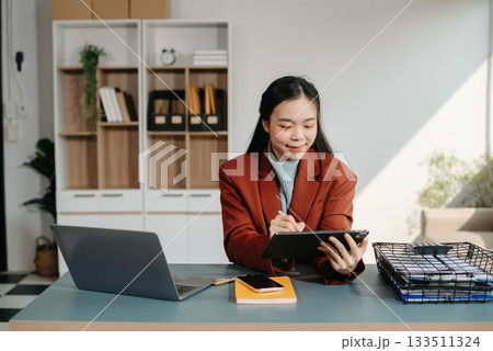 Asian businesswoman working with working notepad, tablet and laptop documents talking on the smartphone, tablet and laptop video call tax, report, accounting 133511324