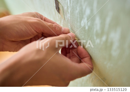 Closeup of textured plaster wall prep, Gently detaching thin tape from wall surface, Homeowner carefully removing adhesive strip to ready wall for repainting process Closeup of textured plaster wall prep, Gently detaching thin tape from wall surface, Homeowner carefully removing adhesive strip to ready wall for repainting process 133515120