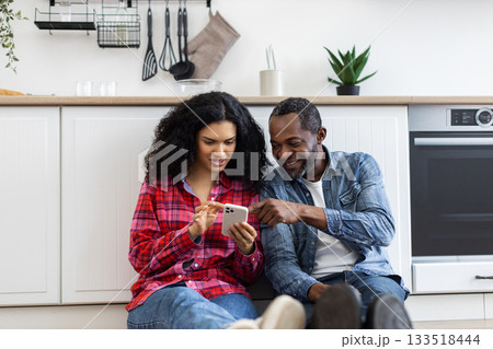 A mixed-race couple sits on the floor in a modern kitchen, looking at a smartphone together. A mixed-race couple sits on the floor in a modern kitchen, looking at a smartphone together. 133518444