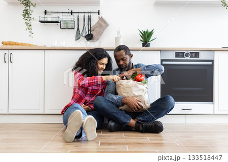 A couple sits on the floor in their kitchen, unpacking a paper grocery bag filled with fresh produce. A couple sits on the floor in their kitchen, unpacking a paper grocery bag filled with fresh produce. 133518447