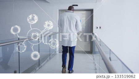Walking doctor wearing lab coat down hospital hallway, with glass railing, digital network overlay 133519062