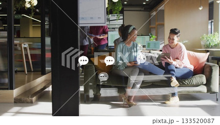 Discussing data two women wearing business attire reviewing tablet in office lounge, with reports 133520087