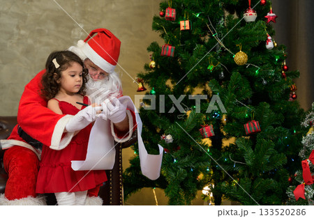 Santa Claus helping young girl write Christmas wish list beside decorated christmas tree in festive Indoor holiday celebration Santa Claus helping young girl write Christmas wish list beside decorated christmas tree in festive Indoor holiday celebration 133520286