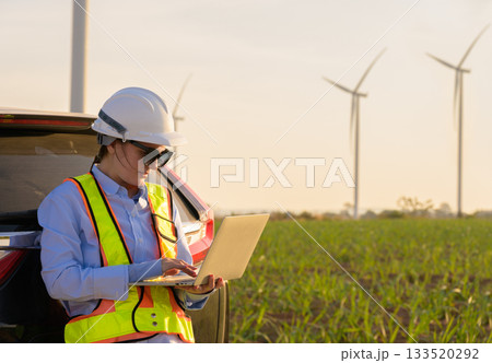 Female engineer in white helmet working on laptop beside vehicle at wind farm. Female wearing high visibility vest and sunglasses monitoring operations with wind turbines in green fields Female engineer in white helmet working on laptop beside vehicle at wind farm. Female wearing high visibility vest and sunglasses monitoring operations with wind turbines in green fields 133520292