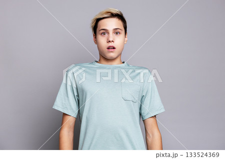 Surprised teenage boy portrait in studio on gray background. Shocked young male model with expressive face and blond hair, showing emotional reaction 133524369