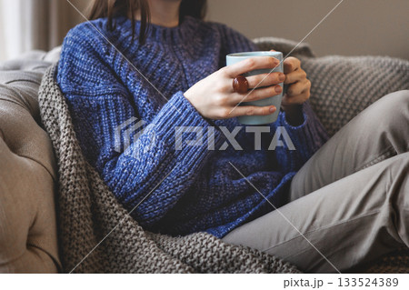 Female hands holding blue mug while sitting in oversized knitted sweater and warm blanket. Cozy autumn or winter atmosphere indoors with comfort Female hands holding blue mug while sitting in oversized knitted sweater and warm blanket. Cozy autumn or winter atmosphere indoors with comfort 133524389