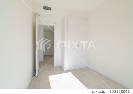 A bright, empty room with white walls and light wooden flooring. A closed door leads to another space, and a wardrobe is visible against the wall. 133528041