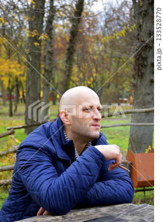 bald young man in a jacket smokes a cigarette on a bench in the autumn park 133528770
