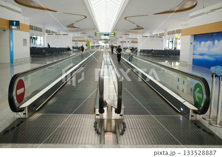 People walking on a moving walkway at a modern airport terminal with seating areas and bright overhead lights during daylight hours People walking on a moving walkway at a modern airport terminal with seating areas and bright overhead lights during daylight hours 133528887