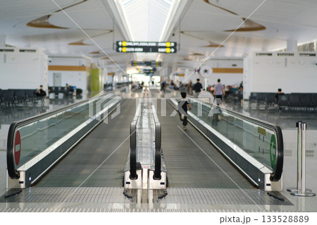 Wide and modern airport terminal with moving walkway and people walking, showcasing a bright and spacious environment during daytime 133528889
