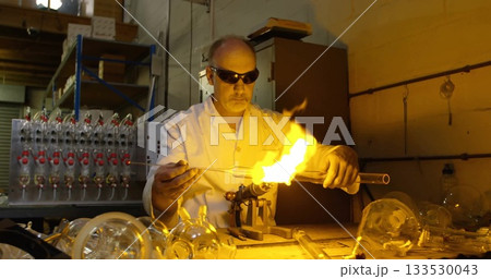 Shaping male glassblower directing flame onto glass tube at studio, with torch and lab coat 133530043