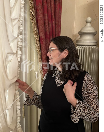 Woman stands by the window, admiring the view through beautiful curtains 133532025