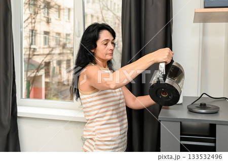 A 50 year old Russian woman with long dark hair pours hot water into a mug for morning coffee, still sleepy, enjoying a calm start to her new day in a cozy kitchen. A 50 year old Russian woman with long dark hair pours hot water into a mug for morning coffee, still sleepy, enjoying a calm start to her new day in a cozy kitchen. 133532496