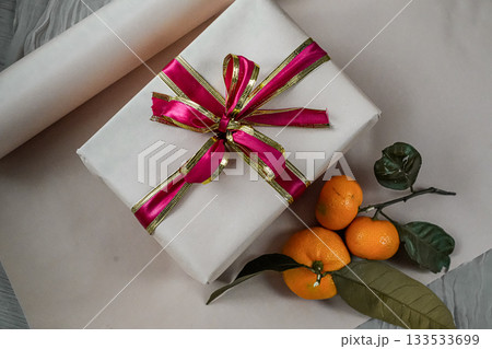 Medium view of a beige gift box decorated with a red ribbon, placed on a neutral background. Two fresh tangerines lie beside it, creating a warm and festive composition suitable for Christmas, New Medium view of a beige gift box decorated with a red ribbon, placed on a neutral background. Two fresh tangerines lie beside it, creating a warm and festive composition suitable for Christmas, New 133533699