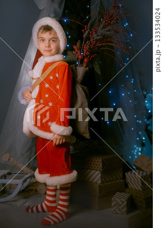 Cute boy dressed as a red and white elf with a backpack, enjoying the Christmas atmosphere near a sparkling decorated tree. 133534024