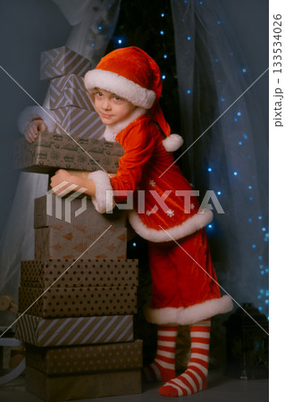 Smiling boy in Santa outfit standing by a Christmas tree with blue lights, embracing a large stack of wrapped gifts Smiling boy in Santa outfit standing by a Christmas tree with blue lights, embracing a large stack of wrapped gifts 133534026