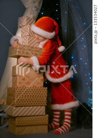 Child dressed as Santa Claus near a festive tree with blue lights, holding many piled-up presents and smiling joyfully Child dressed as Santa Claus near a festive tree with blue lights, holding many piled-up presents and smiling joyfully 133534027