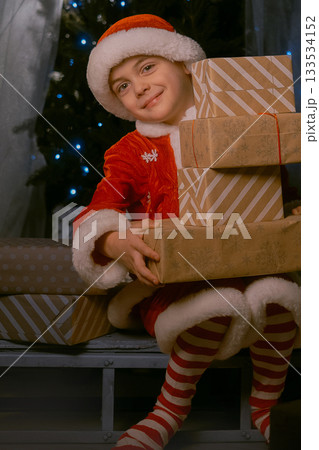 Boy dressed as Santa Claus sitting on sleigh near a Christmas tree with blue lights, holding many wrapped gifts indoors Boy dressed as Santa Claus sitting on sleigh near a Christmas tree with blue lights, holding many wrapped gifts indoors 133534152