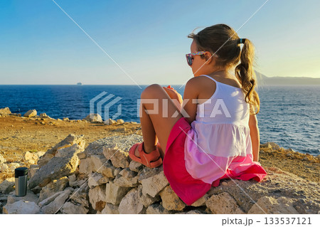 Little 6-year-old girl in striped dress and sunglasses sitting on rocks by the sea 133537121
