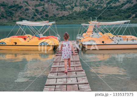 Georgioupoli, Crete, Greece. June 4, 2025. Little girl in pink dress walking on wooden pier to reach catamaran at Lake Kournas 133537375