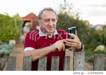 Elderly farmer talking on mobile phone at the fence of his farm on summer evening Elderly farmer talking on mobile phone at the fence of his farm on summer evening 133540045
