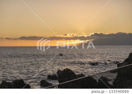 Orange sunset at sea shore promenade in Playa de San Juan, Tenerife, Spain in the south of Tenerife with ocean waves crushing black lava rocks Orange sunset at sea shore promenade in Playa de San Juan, Tenerife, Spain in the south of Tenerife with ocean waves crushing black lava rocks 133540089