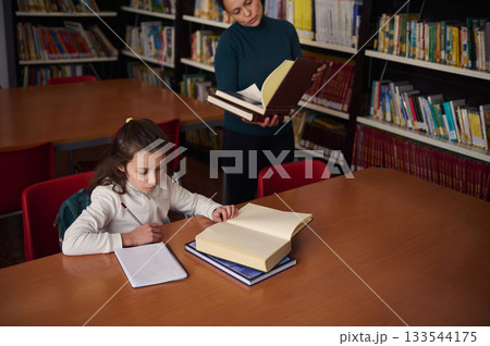 Young Girl Studying in Library While Teacher Holds Books and Guides Learning Young Girl Studying in Library While Teacher Holds Books and Guides Learning 133544175