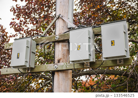 Numbered electrical junction boxes mounted on a wooden utility pole with cables and autumn foliage in the background, part of outdoor power infrastructure. Numbered electrical junction boxes mounted on a wooden utility pole with cables and autumn foliage in the background, part of outdoor power infrastructure. 133546525