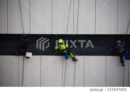 Industrial climbers wipe down building facade element with cloth. Climbers work at height. Safety equipment is essential for dangerous, life threatening work Industrial climbers wipe down building facade element with cloth. Climbers work at height. Safety equipment is essential for dangerous, life threatening work 133547805