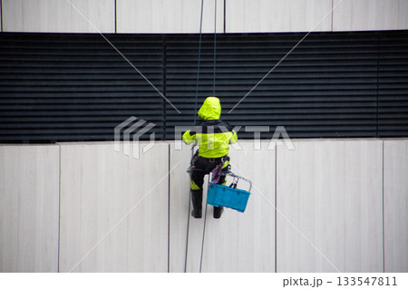 Industrial climber in reflective construction overalls sits on a special seat for working at height. Dangerous work and poor weather conditions. Industrial mountaineering as risky profession 133547811