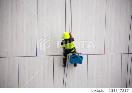 Industrial climber in bright suit descends using safety ropes after completing work at height. The industrial climber work is complete. Ending the workday at height Industrial climber in bright suit descends using safety ropes after completing work at height. The industrial climber work is complete. Ending the workday at height 133547817