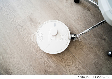 Robotic vacuum navigating near office chair wheel on wooden flooring during automated cleaning routine at home 133548237