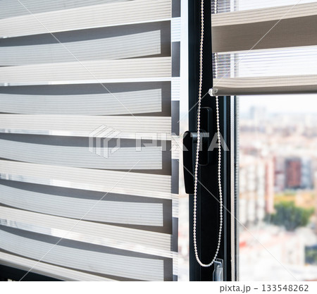 Close-up of zebra roller blinds with window handle and chain mechanism against bright city background 133548262