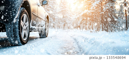 A car is parked on a snow-covered road, surrounded by trees. Snow falls gently, illuminated by warm sunlight in the distance 133548694