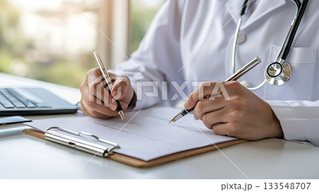 A healthcare worker is focused on writing notes and reviewing documents using two pens at a well-lit desk with a laptop A healthcare worker is focused on writing notes and reviewing documents using two pens at a well-lit desk with a laptop 133548707