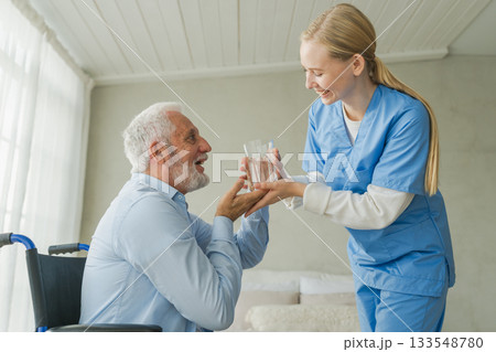 Help support healthcare water balance. Nurse giving glass water for old man patient in wheelchair. Paralyzed man in chair for people with disability drinking fresh water. Senior man and nurse at home 133548780